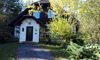 A charming house with a white facade and a black roof, surrounded by trees and a garden. The entrance is invitingly designed, with a paved path.