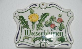 A decorative ceramic sign with the inscription "Meadow Flowers". It features various flowers, including dandelions and clover, in vibrant colors.