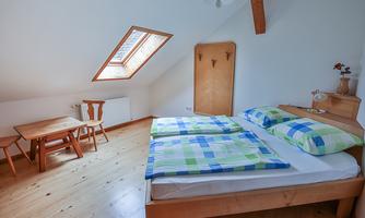 A cozy bedroom with a double bed and colorful blankets. A small table with chairs and a window provide plenty of daylight.