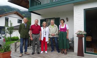 A group of five people is standing in front of a house. They are wearing different styles of clothing, including traditional costumes.