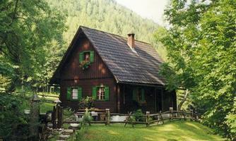 A charming wooden house surrounded by trees and a green property. The windows are adorned with flowers and there is a small pathway to the entrance.