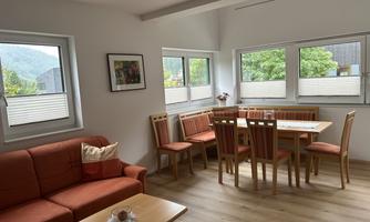 A bright living room with a red sofa and a wooden table. Large windows offer a view of the green surroundings.