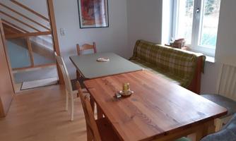A cozy dining area with a wooden table, a green table, and a couch. Large windows provide plenty of daylight in the room.