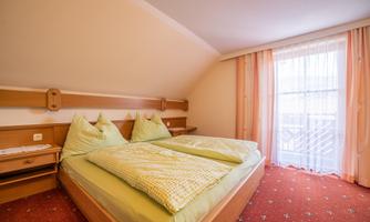 A cozy bedroom with a double bed and green bedspreads. The room has large windows with light curtains and a warm carpet floor.