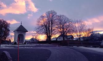 A picturesque winter landscape with a small pavilion and trees in front of a colorful sunset. The sky displays a beautiful play of colors in pink and purple.