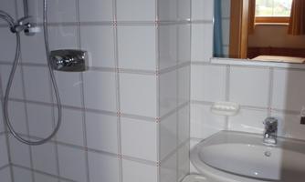 A bright bathroom with white tiles, a shower, and a sink. A window allows daylight in and provides a view outside.