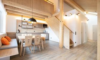 A modern, bright living room with wooden decor and an open kitchen. The room features a dining table and a cozy seating area.
