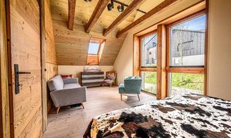 A cozy bedroom with a wooden ceiling and large windows. It includes a bed, a desk, and comfortable seating.