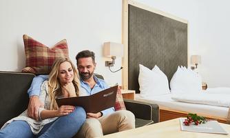 A happy couple is sitting on a couch, looking at a menu. In the background, you can see a cozy room with a bed and modern furnishings.
