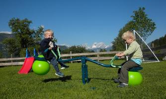 Two children are playing on a seesaw outdoors. In the background, green meadows and snow-covered mountains can be seen.