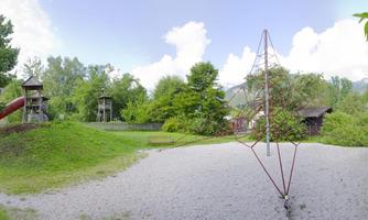 A playground with a slide and several climbing frames is surrounded by trees and green areas. The ground is covered with sand and gravel.
