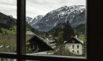 View from a window onto a picturesque mountain landscape with snow-covered peaks. In the foreground are traditional houses and plenty of greenery.
