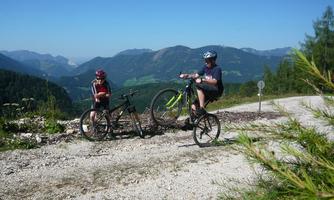 Two children are riding their bikes on a narrow path in the mountains. In the background, green valleys and mountain landscapes can be seen.
