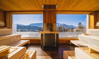 A modern sauna interior with wooden benches and large windows. The view extends to the mountains and the blue sky.