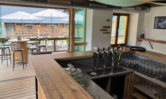 A modern designed bar with a wooden counter and several taps. In the background, there are tables and chairs on a terrace overlooking the outdoors.