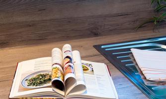 An open cookbook lies on a table. Next to the book is a sink and a kitchen towel.