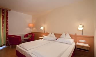 A cozy hotel room with two single beds and red accents. The room is well-lit and features an armchair as well as bedside tables.