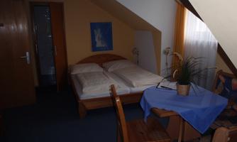 A cozy room with a double bed and a table. The walls are painted in warm colors and there is a view of the room from a window.