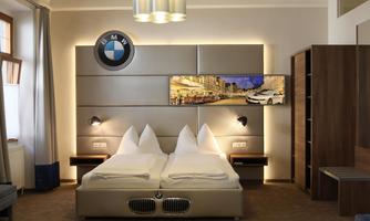 A modern hotel room with a BMW theme. The bed has a sleek design with BMW elements and a TV wall with a city view.