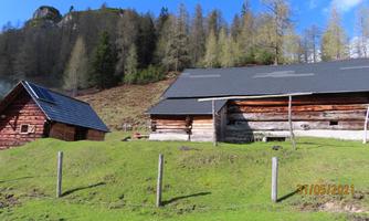 A landscape picture with two wooden sheds on a green meadow. In the background, trees and gentle hills are visible.