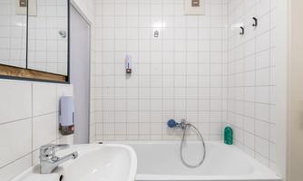 A simple bathroom with a bathtub and a sink. The walls are tiled, and there is a mirror as well as corresponding soap dispensers.