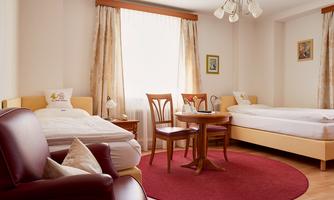 A cozy hotel room with two beds, a red carpet, and a small table. Bright windows create a friendly atmosphere.