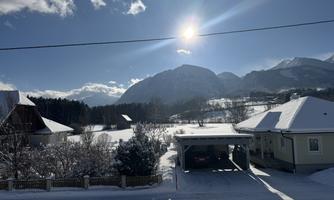 A winter landscape with snow-covered mountains and radiant sunshine. In the foreground, there are some houses and trees visible.