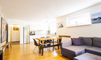 A modern living room with a large couch and a dining table. The bright walls and wooden floor create a friendly atmosphere.
