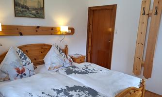A cozy bedroom with a double bed and wooden furniture. Two lamps and a small picture hang on the wall, while a door leads into the background.