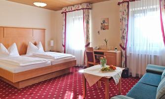 A bright hotel room with a double bed, a desk, and a cozy sofa. Large windows provide plenty of daylight and a friendly atmosphere.