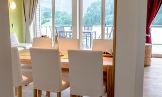 A bright dining area with a large table and white chairs. The windows offer a beautiful view of the surrounding nature.