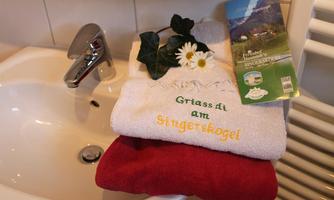 A sink with a towel in white and red as well as a greeting card. The towel is embroidered with "Grüß di am Singerskogel" and decorated with a plant and a flower.
