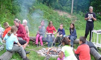 A group of children is seated around a campfire, roasting sausages. In the background, trees and a green meadow can be seen.