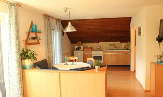 A bright and inviting living kitchen with a wooden beam ceiling. The room is simply furnished, with a dining table and an open kitchenette.