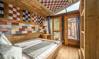 A cozy bedroom with colorful walls and wooden paneling. There is a large window that lets in plenty of daylight.