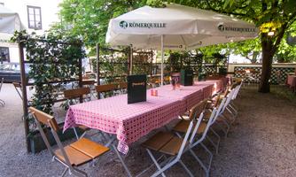 A cozy outdoor area with a long table and chairs. A parasol provides shade in a green garden.