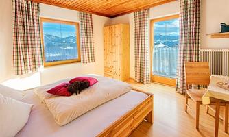 A cozy bedroom with wooden decor and large windows. The view shows a beautiful landscape with mountains and a clear sky.