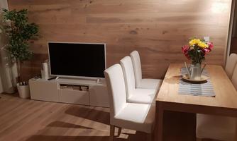 A modern furnished living space with a table and white chairs. On the table, there is a vase of flowers and a television is visible in the background.