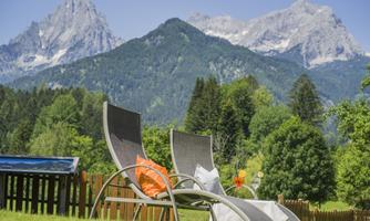 Two lounge chairs are in a green meadow with mountains in the background. The sky is clear, and nature appears inviting.