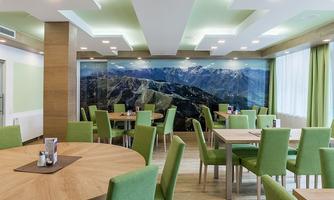 A modern restaurant with round wooden tables and green chairs. The wall features an impressive mountain panorama and large windows provide plenty of daylight.
