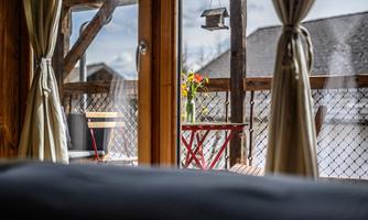 A cozy balcony with a red table and a chair. In the background, flowers in a vase and a beautiful view are visible.