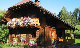 A charming wooden house with a balcony, adorned with colorful flowers. Surrounded by green countryside and trees.