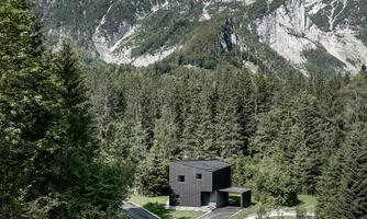 A modern black house stands amidst a wooded landscape. Majestic mountains rise in the background under a clear sky.