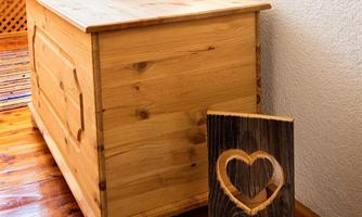 A wooden trunk furniture piece with an orange cushion on top and a decorative wooden panel with a heart motif next to it. The floor is made of wood, creating a cozy atmosphere.