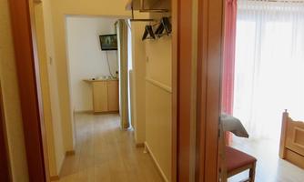 A bright hallway with a wooden floor and coat rack. In the background, you can see a television and a chair next to a window.