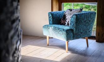 A stylish armchair in turquoise with a decorative cushion stands in front of a window, letting in daylight. The wooden floor enhances the cozy atmosphere of the room.