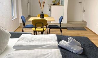 A modern room with a large bed and fresh towels. In the background, there is a dining area with a table and chairs.