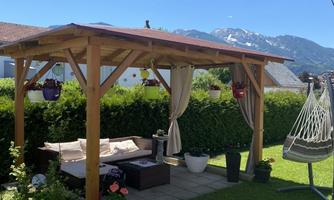 A cozy pavilion in the garden with seating and plants. In the background, the mountains and a clear blue sky can be seen.