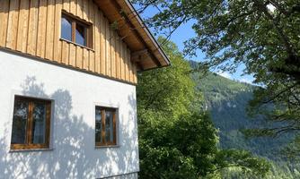 A charming house with wooden cladding and white walls, surrounded by green trees. In the background, picturesque mountains stretch out.