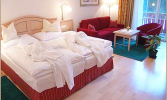 A cozy hotel room with a large bed and fresh sheets. The room features a red sofa, a table, and a plant.